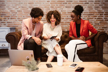 Gruppe von fröhlichen multiethnischen weiblichen Mitarbeitern in formeller Kleidung, die sich Notizen in einem Notizbuch machen, während sie zusammen auf einer Couch in einem hellen Büro arbeiten - ADSF34086