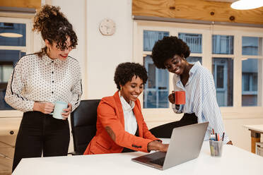 Gruppe glücklicher multiethnischer weiblicher Mitarbeiter in eleganter Kleidung beim Surfen auf einem Netbook, während sie gemeinsam an einem Projekt in einem hellen, modernen Büro arbeiten - ADSF34087