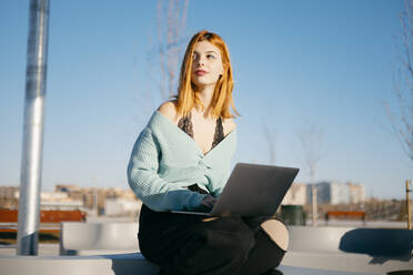 Attraktive Frau mit roten Haaren surft auf einem modernen Netbook, während sie auf einer Straße mit Wohngebäuden an einem sonnigen Tag in der Stadt sitzt und wegschaut - ADSF34090