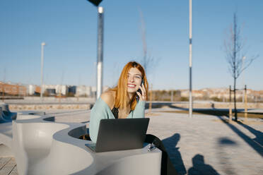 Lächelnde Frau, die wegschaut und mit ihrem Smartphone spricht, während sie in der Nähe eines modernen Netbooks auf der Straße einer Stadt mit hellem Sonnenlicht sitzt - ADSF34092