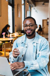 Lächelnder afroamerikanischer Mann mit einer Tasse Kaffee, der in die Kamera schaut, während er am Tisch in einem modernen Café in der Pause auf seinem Netbook surft - ADSF34097