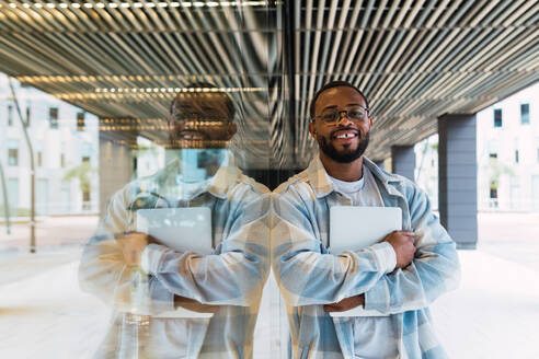 Lächelnder afroamerikanischer Mann mit modernem Netbook in den Händen, der in die Kamera schaut, während er an der Glaswand eines modernen Gebäudes auf der Straße steht - ADSF34099