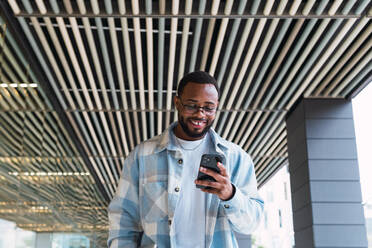Positiver afroamerikanischer Mann mit Brille, der mit seinem Handy SMS schreibt, während er in der Nähe eines modernen Gebäudes mit Glaswand auf der Straße steht - ADSF34100