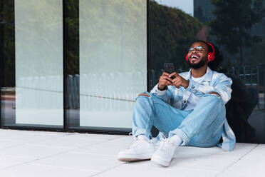 Ganzkörperaufnahme eines glücklichen afroamerikanischen Mannes mit Handy, der mit drahtlosen Kopfhörern Musik hört, während er an der Wand eines modernen Gebäudes sitzt - ADSF34101