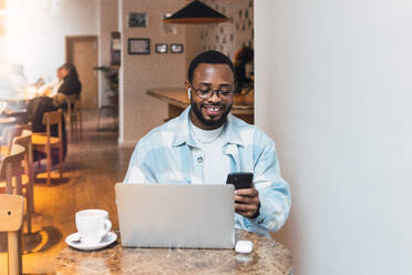 Lächelnder afroamerikanischer Mann mit drahtlosen Kopfhörern, der mit seinem Handy Textnachrichten sendet, während er mit seinem Laptop in einer modernen Cafeteria am Tisch sitzt - ADSF34102