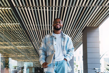 From below smiling African American male with modern netbook in hands looking away while walking near glass wall of modern building on street - ADSF34103