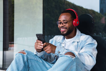Glücklicher afroamerikanischer Mann mit Mobiltelefon, der mit drahtlosen Kopfhörern Musik hört, während er an der Wand eines modernen Gebäudes sitzt - ADSF34104