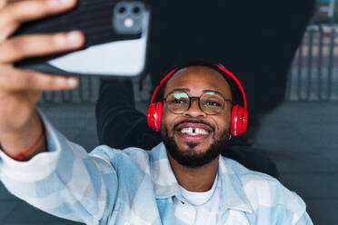 From above of positive African American male in wireless red headphones taking self portrait on smartphone while standing on street - ADSF34106