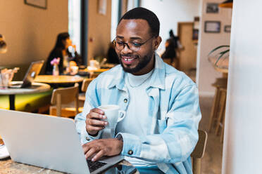 Lächelnder afroamerikanischer Mann mit einer Tasse Kaffee, der am Tisch in einem modernen Café in der Pause auf seinem Netbook surft - ADSF34108