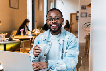 Lächelnder afroamerikanischer Mann mit einer Tasse Kaffee, der wegschaut, während er am Tisch in einem modernen Café in der Pause auf seinem Netbook surft - ADSF34109