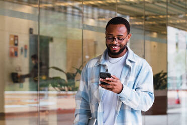 Positiver afroamerikanischer Mann in modischer Kleidung und mit Brille, der mit seinem Handy SMS schreibt, während er in der Nähe eines Gebäudes mit Glaswand steht - ADSF34110