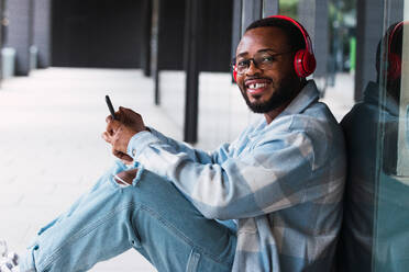 Glücklicher afroamerikanischer Mann mit Mobiltelefon, der mit drahtlosen Kopfhörern Musik hört, während er sitzend in die Kamera auf die Wand eines modernen Gebäudes schaut - ADSF34111