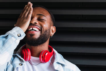 Cheerful bearded African American male with modern red wireless headphones covering closed eye while standing on black background on street - ADSF34112