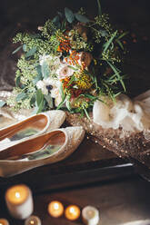 High angle of tender blooms with green leaves arranged in bouquet and placed against bridal shoes and blurred burning candles - ADSF34114