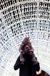 Glücklicher Vater, der seine Tochter auf den Schultern trägt, steht an Weihnachten in einem beleuchteten Tunnel - GMLF01275
