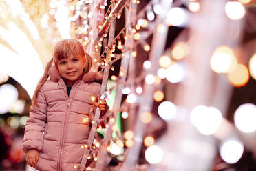 Lächelndes süßes Mädchen, das vor Weihnachtsbeleuchtung steht - GMLF01281