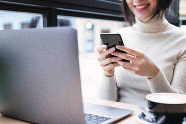 Smiling businesswoman with laptop using mobile phone sitting at cafe - PNAF03409