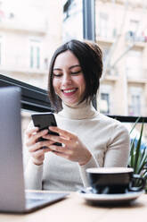 Glückliche Geschäftsfrau mit Laptop und Mobiltelefon in einem Café sitzend - PNAF03410