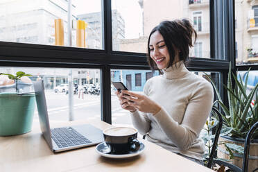 Glückliche Geschäftsfrau mit Laptop und Smartphone in einem Café sitzend - PNAF03413