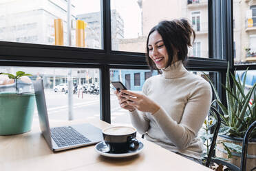 Happy businesswoman with laptop using smart phone sitting in cafe - PNAF03413