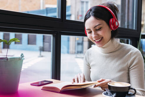 Lächelnde Frau, die über drahtlose Kopfhörer ein Lied hört und ein Buch liest, während sie in einem Café sitzt - PNAF03414