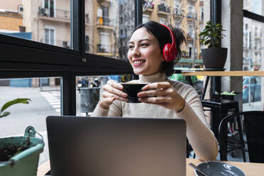 Lächelnde Geschäftsfrau, die eine Kaffeetasse hält und über Kopfhörer in einem Café Musik hört - PNAF03415