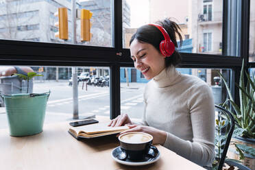 Glückliche Frau, die ein Lied über drahtlose Kopfhörer hört und ein Buch liest, während sie in einem Café sitzt - PNAF03416