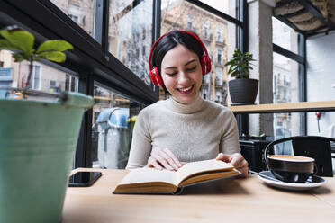 Glückliche Frau mit drahtlosen Kopfhörern, die in einem Café sitzt und ein Buch liest - PNAF03417