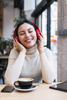 Glückliche Frau mit geschlossenen Augen hört Musik über drahtlose Kopfhörer im Café - PNAF03421