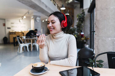 Smiling woman wearing headphones sending voicemail through smart phone at cafe - PNAF03423