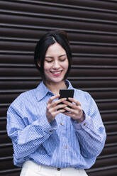 Smiling woman using smart phone standing in front of shutter - PNAF03425