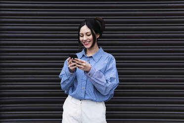 Happy woman using mobile phone standing in front of shutter - PNAF03426