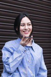 Lächelnde Frau, die über einen Lautsprecher mit ihrem Smartphone spricht und vor einem Fensterladen steht - PNAF03427