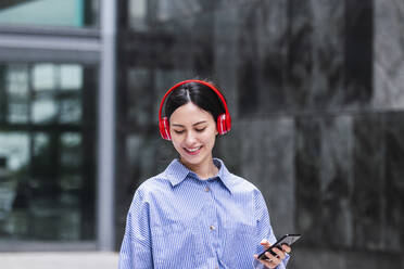 Lächelnde Frau, die ein Smartphone hält und über drahtlose Kopfhörer Musik hört, an einem sonnigen Tag - PNAF03429
