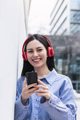 Lächelnde Frau mit Handy in der Hand hört Musik über drahtlose Kopfhörer - PNAF03432