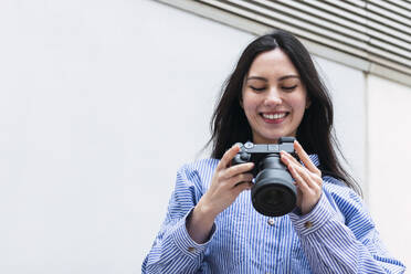 Happy woman holding camera standing in front of white wall - PNAF03439