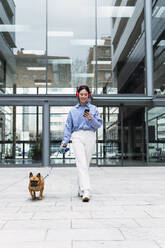 Happy woman listening music through wireless headphones walking with dog in front of building - PNAF03447