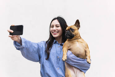 Lächelnde Frau mit Hund, die ein Selfie vor einer weißen Wand macht - PNAF03450