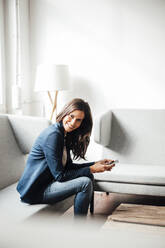 Smiling businesswoman holding smart phone sitting on sofa in office - JOSEF07662