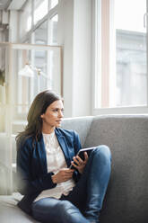 Businesswoman holding tablet PC sitting on sofa looking through window - JOSEF07665