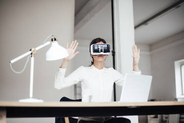 Businesswoman wearing virtual reality simulator gesturing sitting at desk in office - JOSEF07676