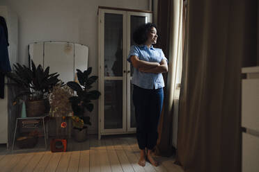 Smiling woman standing with arms crossed looking out through window at home - JOSEF07693