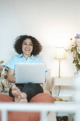 Smiling woman with laptop sitting on bed at home - JOSEF07743