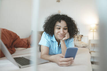 Smiling woman with laptop using smart phone on bed at home - JOSEF07747