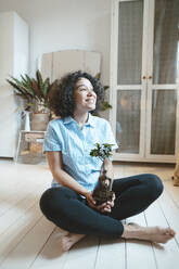 Smiling woman sitting with houseplant on floor at home - JOSEF07750