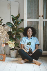 Smiling woman holding houseplant sitting cross-legged on floorboard at home - JOSEF07752