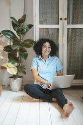 Smiling woman with laptop sitting on floorboard at home - JOSEF07755