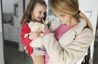Mother carrying smiling daughter holding stuffed toy at dental clinic - JCCMF05799