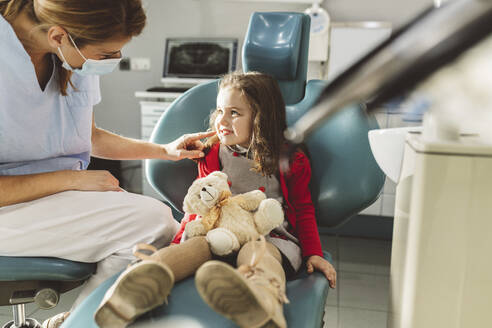 Dentist in protective face mask looking at girl sitting with stuffed toy in dental clinic - JCCMF05819