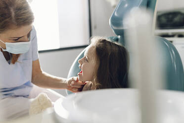 Dentist wearing protective face mask examining teeth of little girl at clinic - JCCMF05821